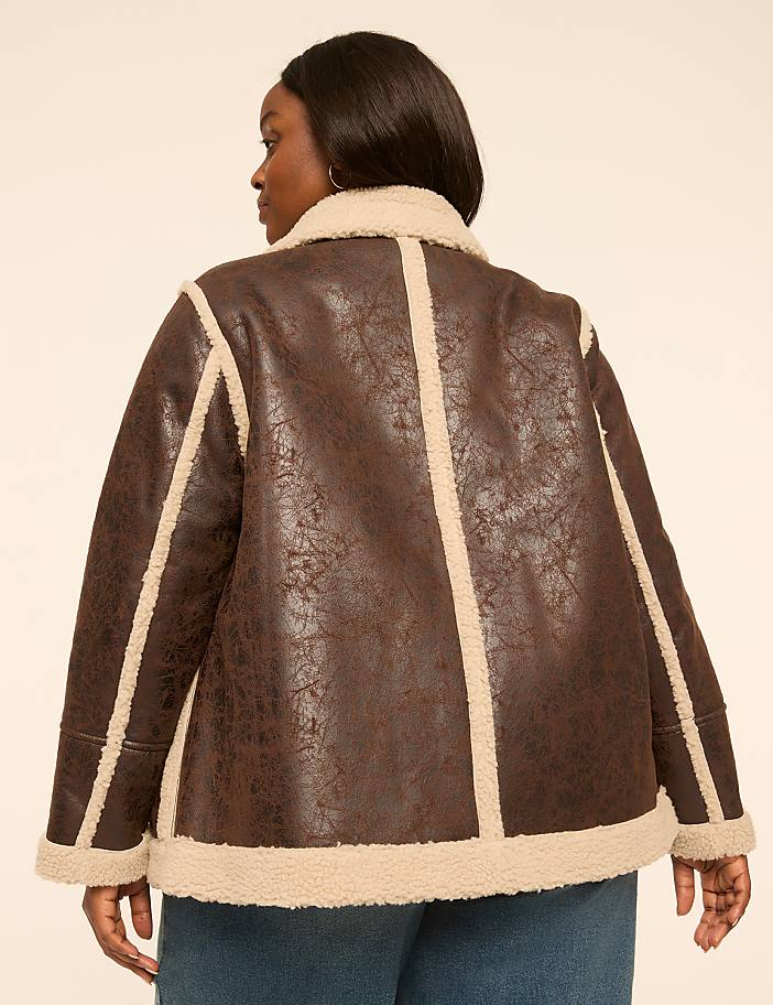A woman wearing a brown faux leather aviator jacket with cream shearling trim stands against a plain, light-colored background.