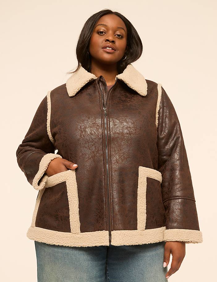 A woman in a brown faux leather aviator jacket with cream shearling trim stands against a plain, light-colored background.
