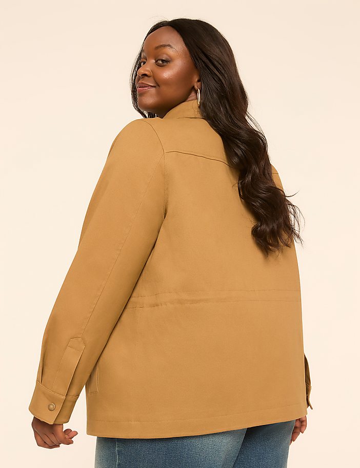 A woman in a tan jacket and blue jeans smiles over her shoulder against a plain, light-colored background.