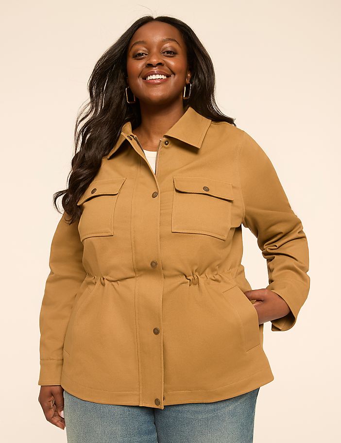 A smiling woman in a tan jacket stands against a plain, light-colored background.