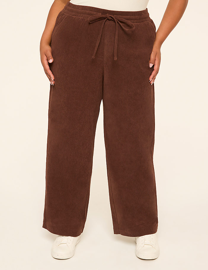 A person stands in the foreground wearing brown corduroy pants and white sneakers against a plain, light-colored background.