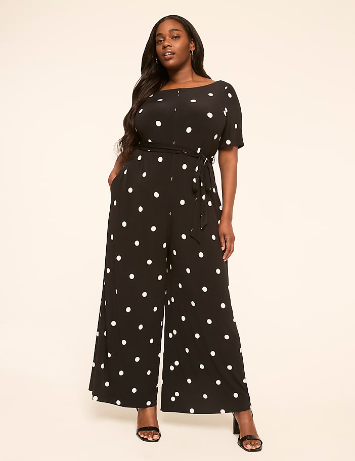 A woman in a black and white polka dot jumpsuit stands against a plain, light-colored background.