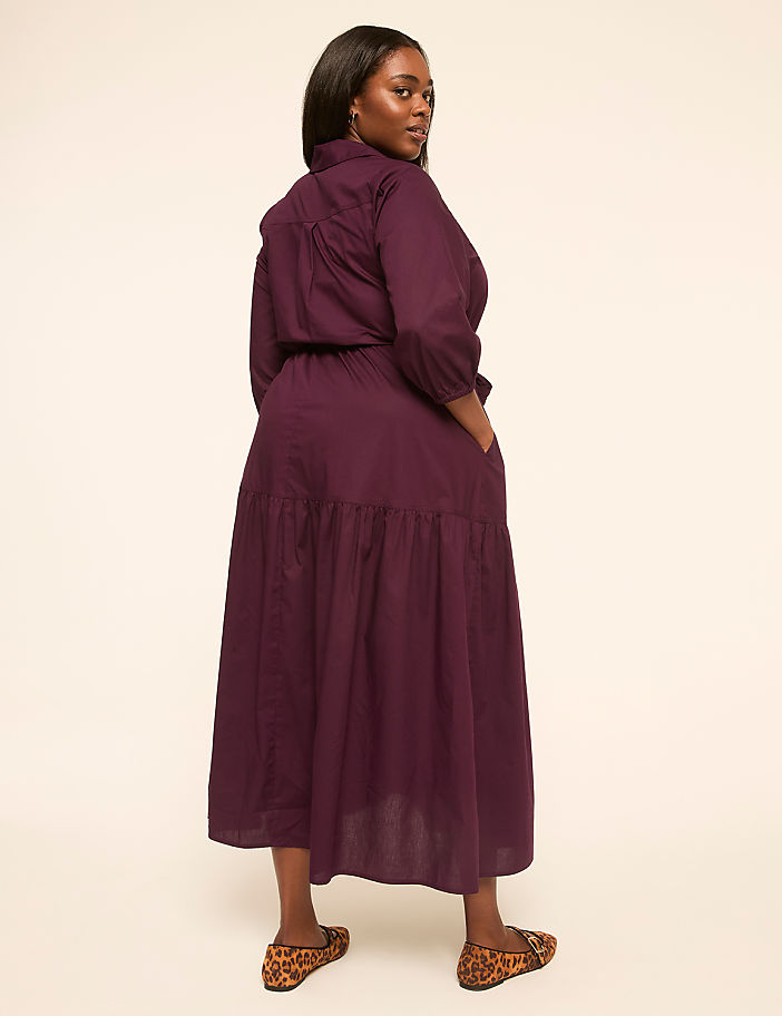 A woman in a purple dress stands against a plain, light-colored background.