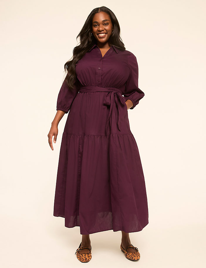 A smiling woman in a plum-colored tiered dress stands against a plain, light-colored background.
