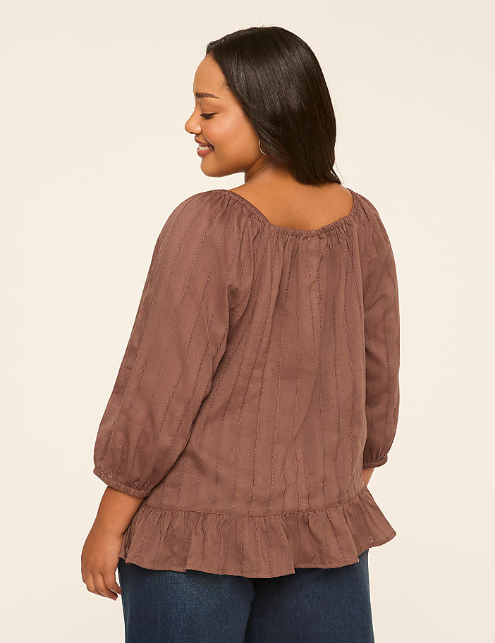 A woman in a brown peasant blouse and blue jeans stands against a plain, light-colored background.