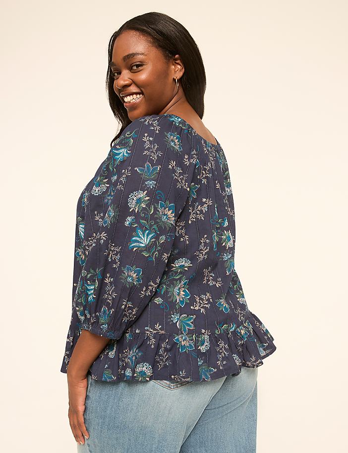 A smiling woman in a floral peplum top and jeans poses against a plain, light-colored background.