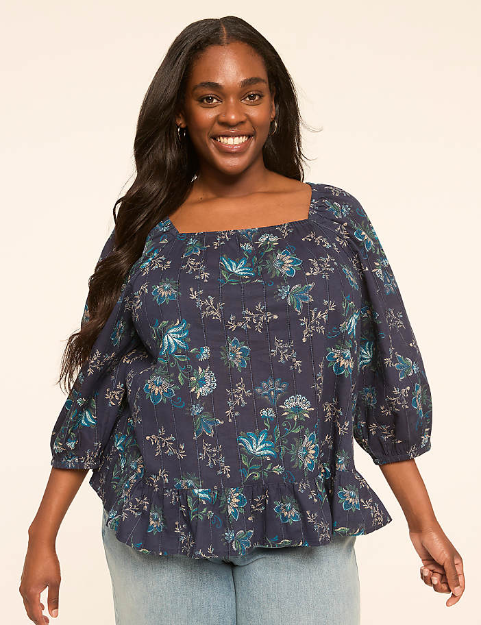 A smiling woman with long dark hair wears a dark blue floral print blouse with a ruffled hem, standing against a plain, light-colored background.