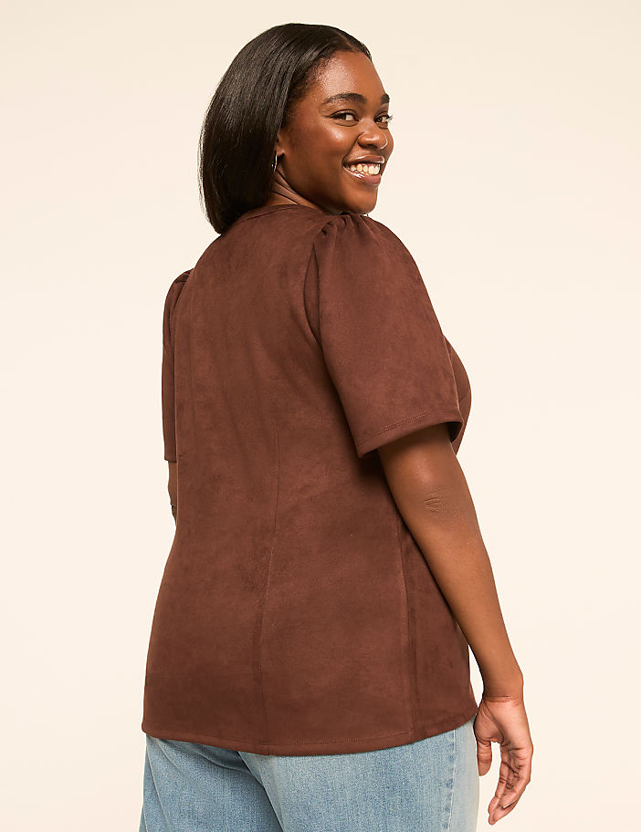 A smiling woman in a brown top and jeans is shown against a plain, light-colored background.