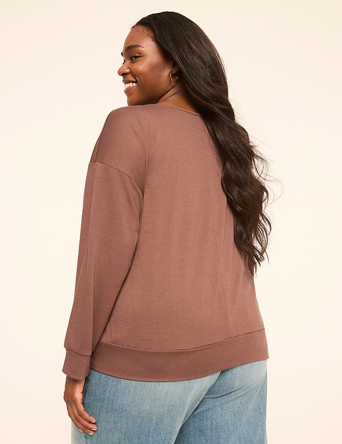 A woman in a brown sweatshirt and blue jeans smiles over her shoulder against a plain, light-colored background.