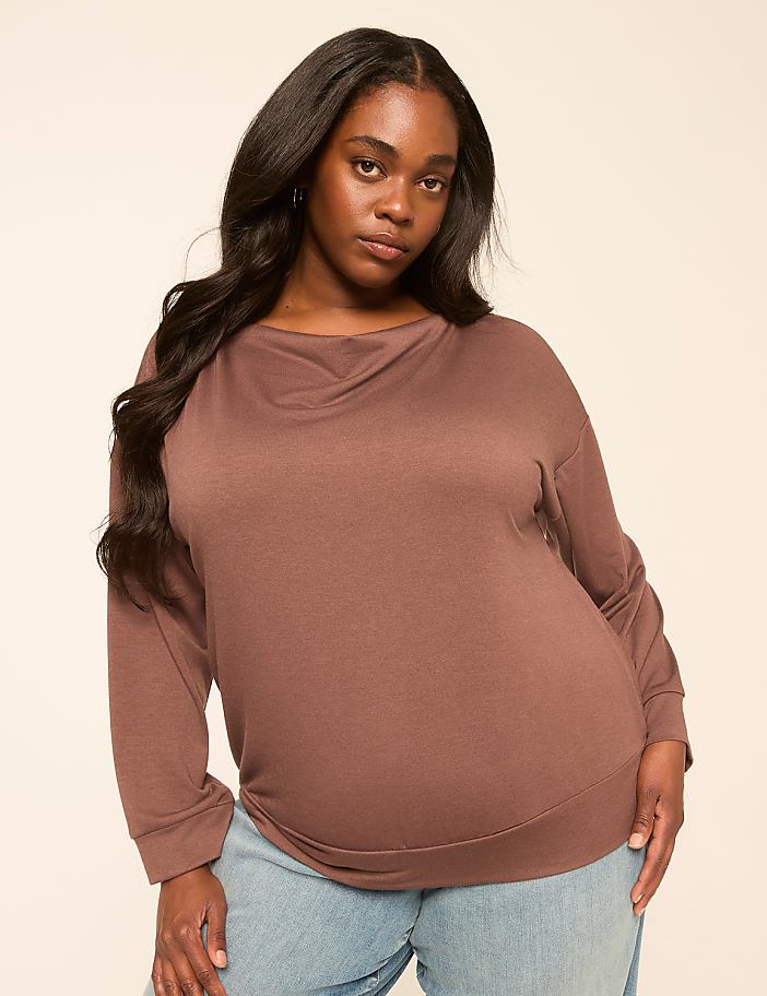 A woman with long, dark hair and a brown long-sleeved shirt stands against a plain, light-colored background.