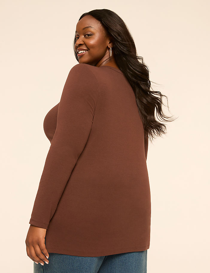 A woman in a brown long-sleeved shirt and blue jeans smiles over her shoulder against a plain, light-colored background.