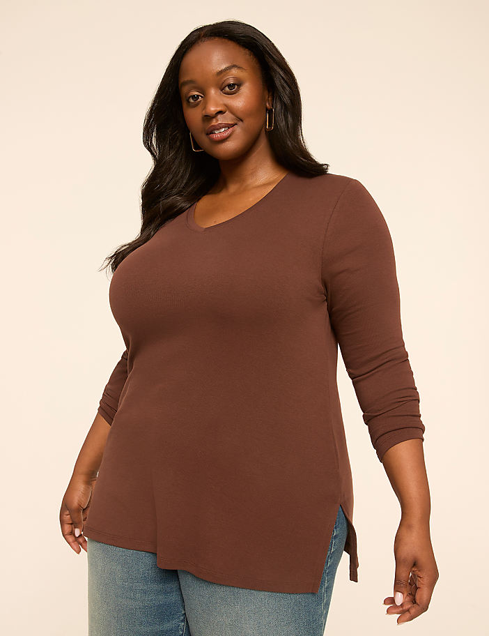 A smiling woman with dark hair and brown skin, wearing a brown v-neck shirt and blue jeans, stands against a plain, light brown background.
