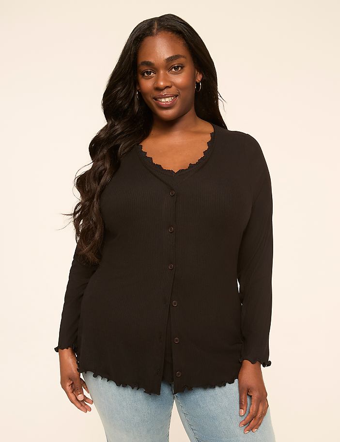A smiling woman with long dark hair wears a black ribbed top with a scalloped neckline and hem, standing against a plain, light-colored background.