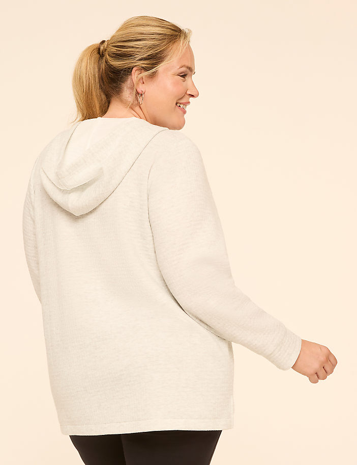 A woman in a light-colored hoodie stands against a plain, light-colored background.