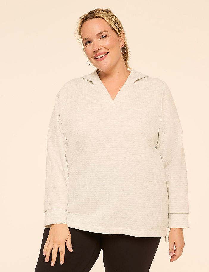 A smiling woman in a light grey hoodie and black leggings stands against a plain, light peach background.