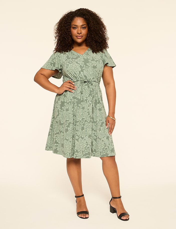 A woman in a green floral dress stands against a plain, light-colored background.
