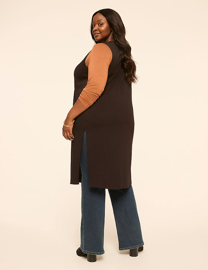 A woman in a long dark vest over a tan long-sleeved shirt and blue jeans stands against a plain, light-colored background.