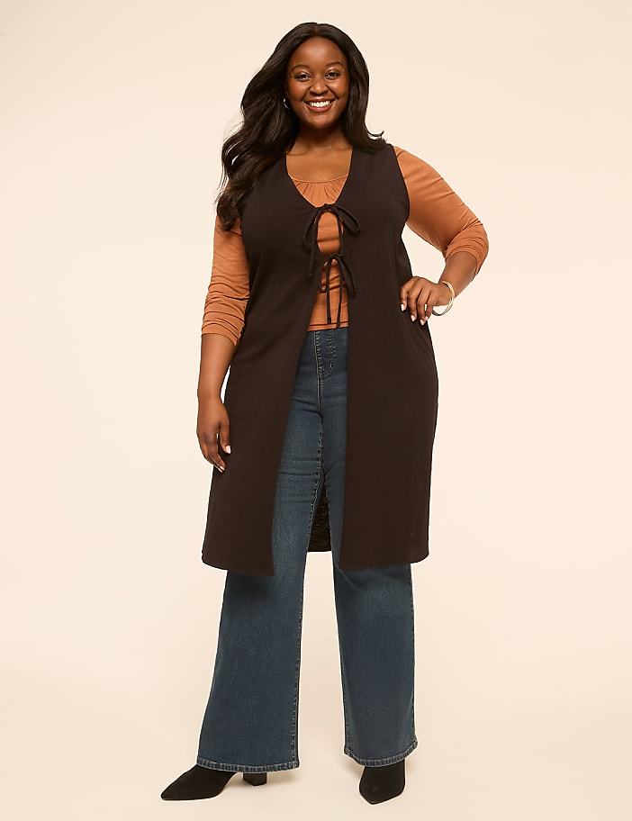 A smiling woman in a long vest and jeans stands against a plain, light-colored background.