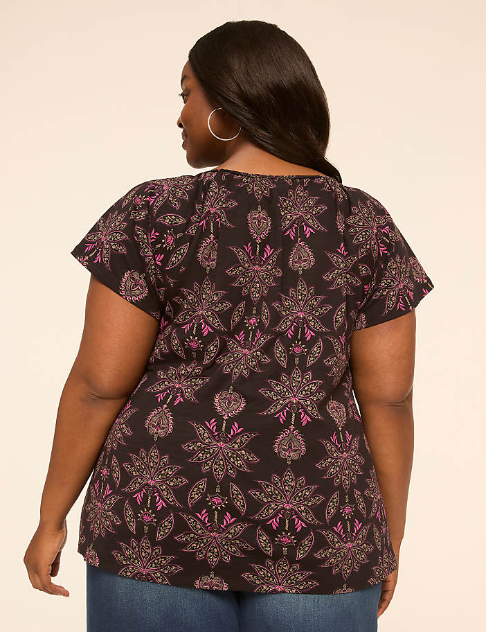 A woman in a patterned top and jeans stands against a plain, light-colored background.