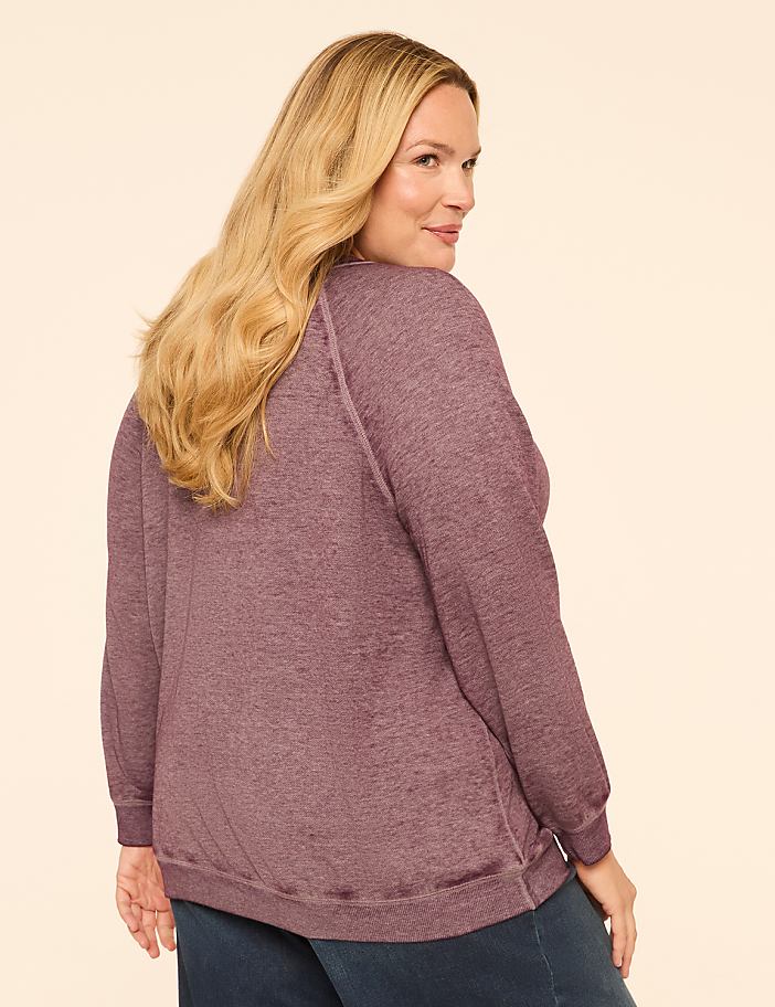 A woman with blonde hair wearing a purple sweatshirt and blue jeans poses against a plain, light-colored background.
