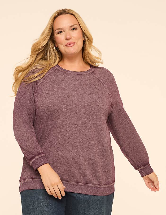 A smiling woman in a maroon sweatshirt and blue jeans stands against a plain, light peach background.