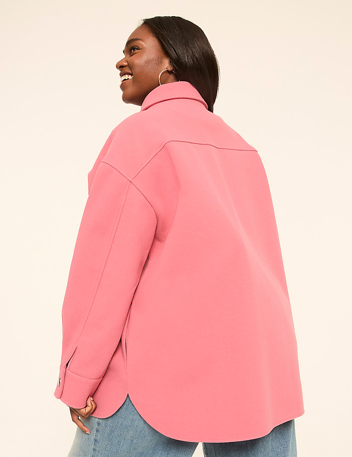 A woman in a pink jacket and jeans smiles over her shoulder against a plain, light background.