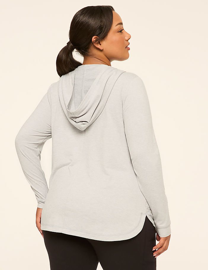 A woman in a light gray hooded shirt and black leggings stands against a plain, light-colored background.