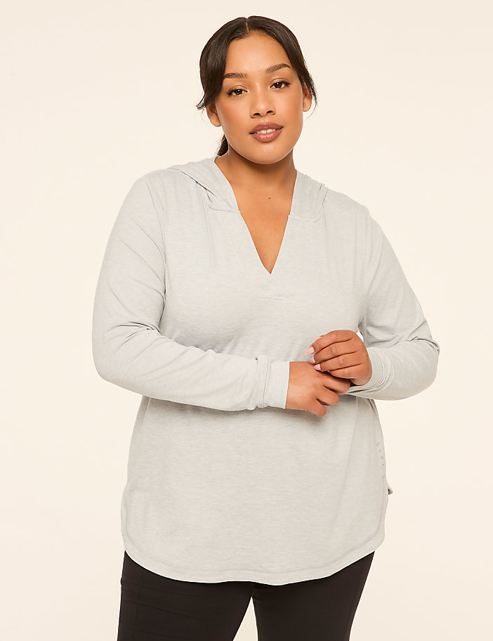 A woman in a light gray hooded shirt stands against a plain, light-colored background.
