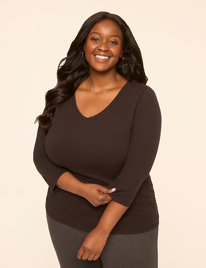 A smiling woman with dark, wavy hair and a black v-neck shirt stands against a plain, light-colored background.