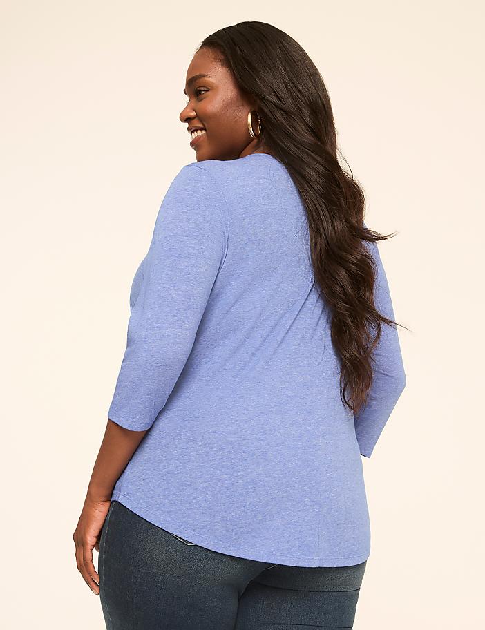 A woman in a blue shirt and jeans stands against a plain, light-colored background.