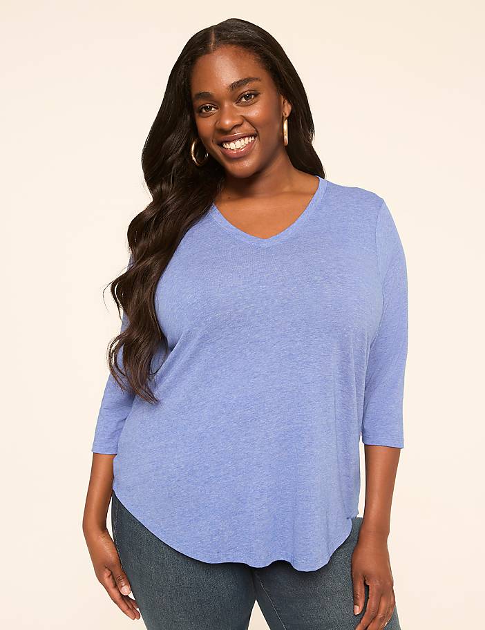 A smiling woman with long dark hair and gold hoop earrings wears a light blue v-neck shirt and jeans against a plain, light-colored background.