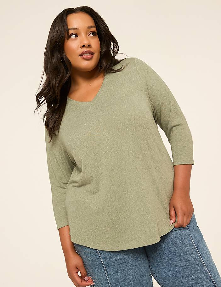 A woman in a green v-neck shirt and blue jeans poses against a plain, light-colored background.