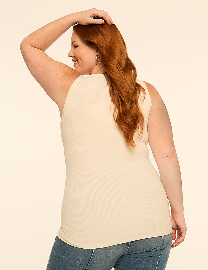 A woman with red hair in a cream-colored ribbed tank top and blue jeans poses against a plain, light-colored background.