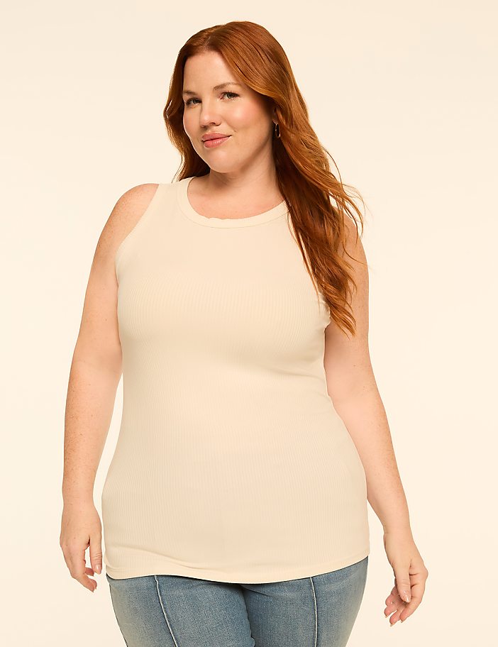 A woman with red hair and a cream-colored ribbed tank top stands against a plain, light-colored background.