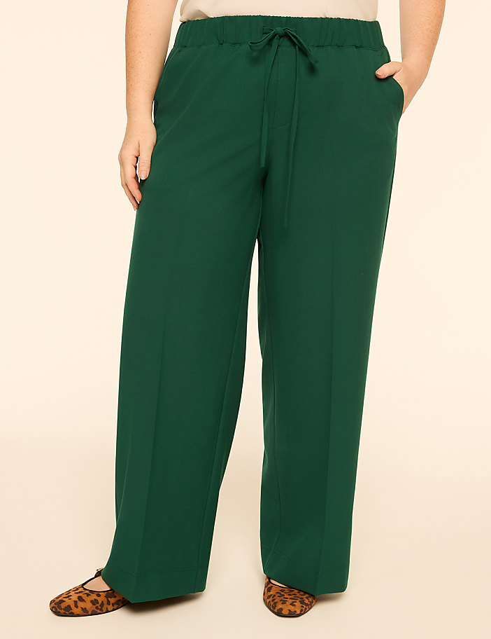 A person stands in wide-leg green pants with leopard print flats against a plain beige background.