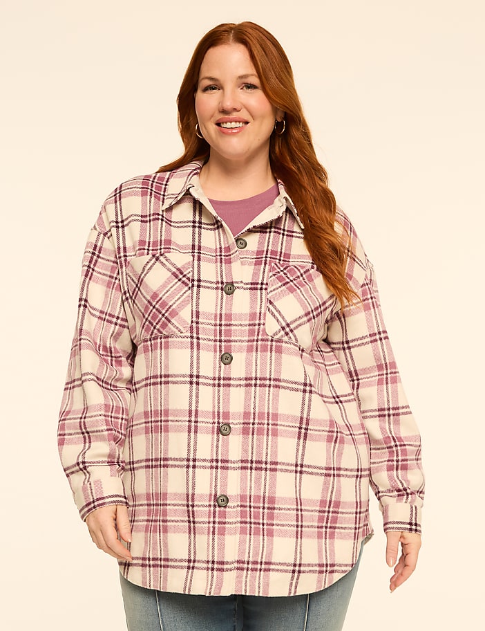 A smiling woman with red hair stands in the foreground wearing a plaid shirt over a purple top, against a plain, light-colored background.
