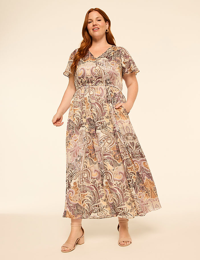 A woman in a flowing paisley dress with flutter sleeves stands with her hands in her pockets against a plain, light-colored background.