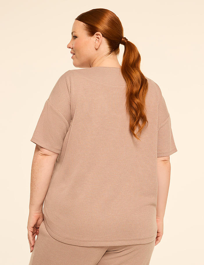 A woman with auburn hair pulled into a ponytail stands facing away from the camera in a beige ribbed t-shirt and matching pants against a plain beige background.