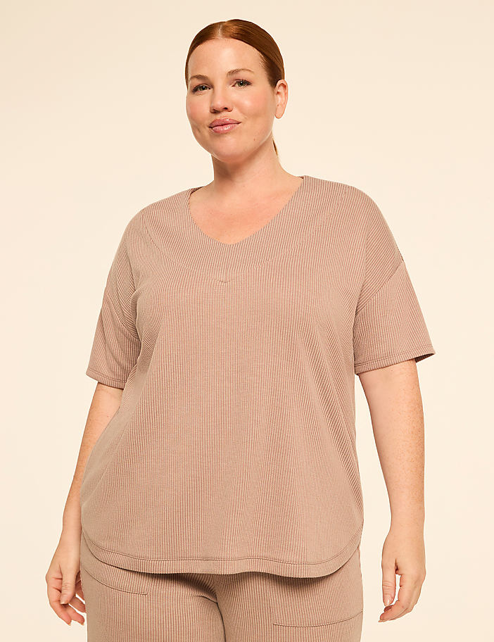 A woman with red hair and fair skin, wearing a beige ribbed v-neck t-shirt and matching pants, stands against a plain, light beige background.
