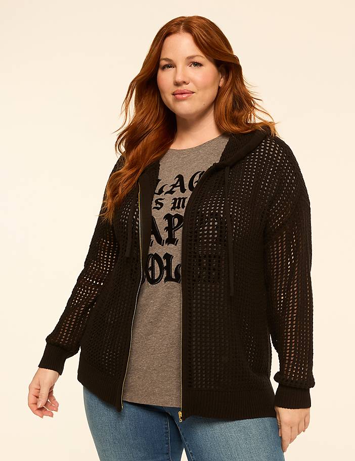 A woman with red hair stands in the foreground wearing a black hooded cardigan over a graphic t-shirt and jeans, against a plain, light-colored background.