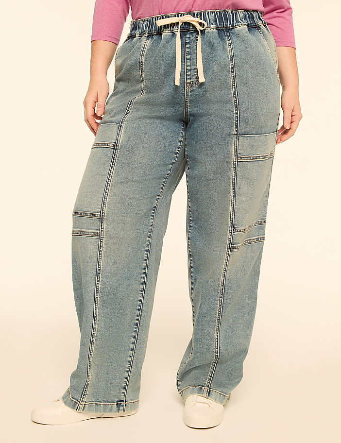 A person stands in light-wash denim cargo pants with a drawstring waist and white sneakers against a plain, light-colored background.