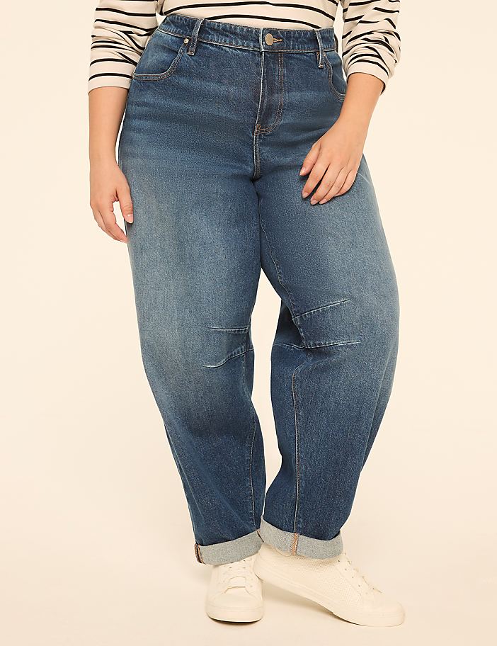 A person wearing baggy blue jeans and white sneakers stands against a plain, light-colored background.
