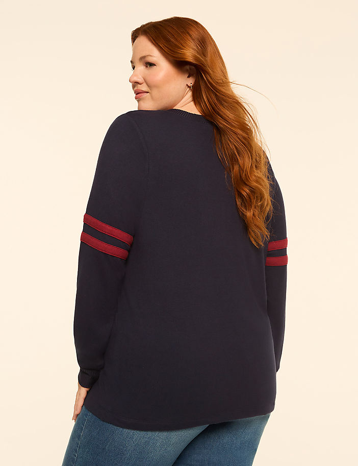A woman with red hair wearing a navy blue long-sleeved shirt with maroon stripes on the sleeves stands against a plain, light-colored background.