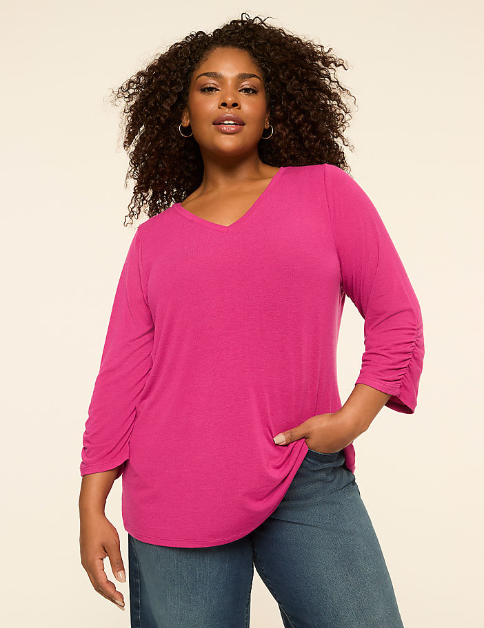A woman in a pink v-neck shirt and blue jeans stands against a plain, light-colored background.