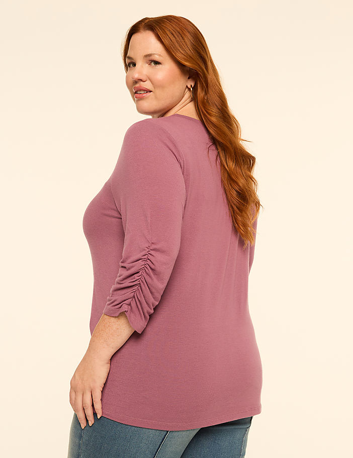 A woman with red hair in a mauve shirt and blue jeans stands against a plain, light-colored background.