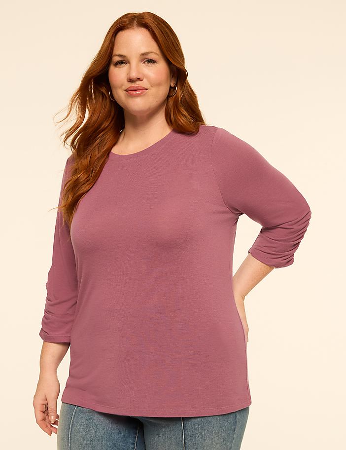 A woman with red hair stands in the foreground wearing a mauve shirt and jeans against a plain, light-colored background.
