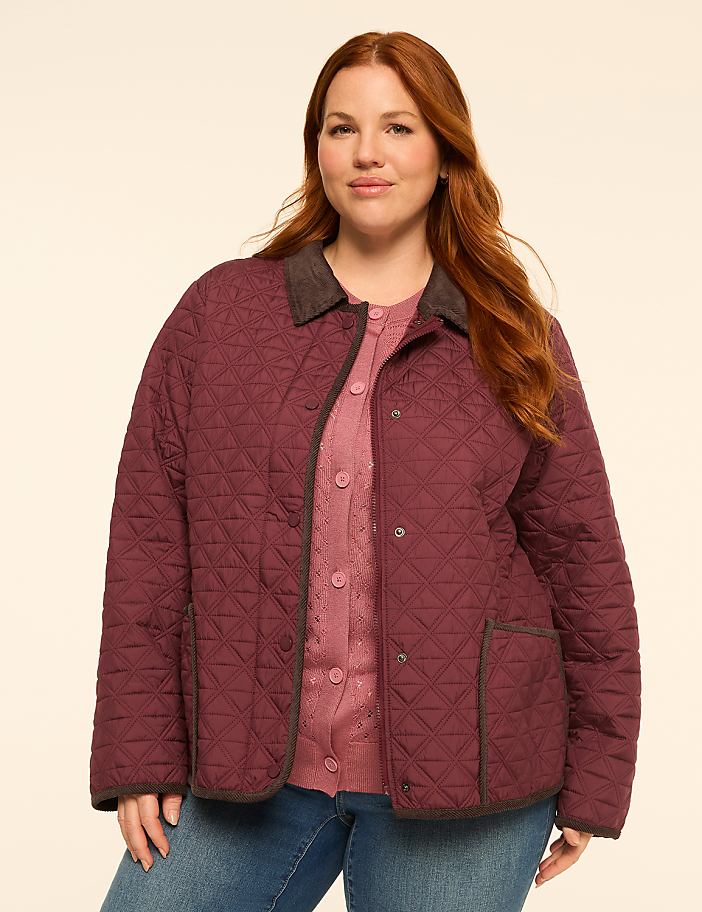 A woman with red hair stands in the foreground wearing a quilted burgundy jacket over a pink cardigan, against a plain, light-colored background.