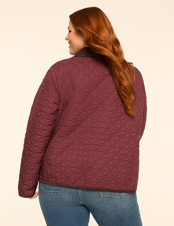 A woman with auburn hair wearing a quilted burgundy jacket and blue jeans stands against a plain, light-colored background.