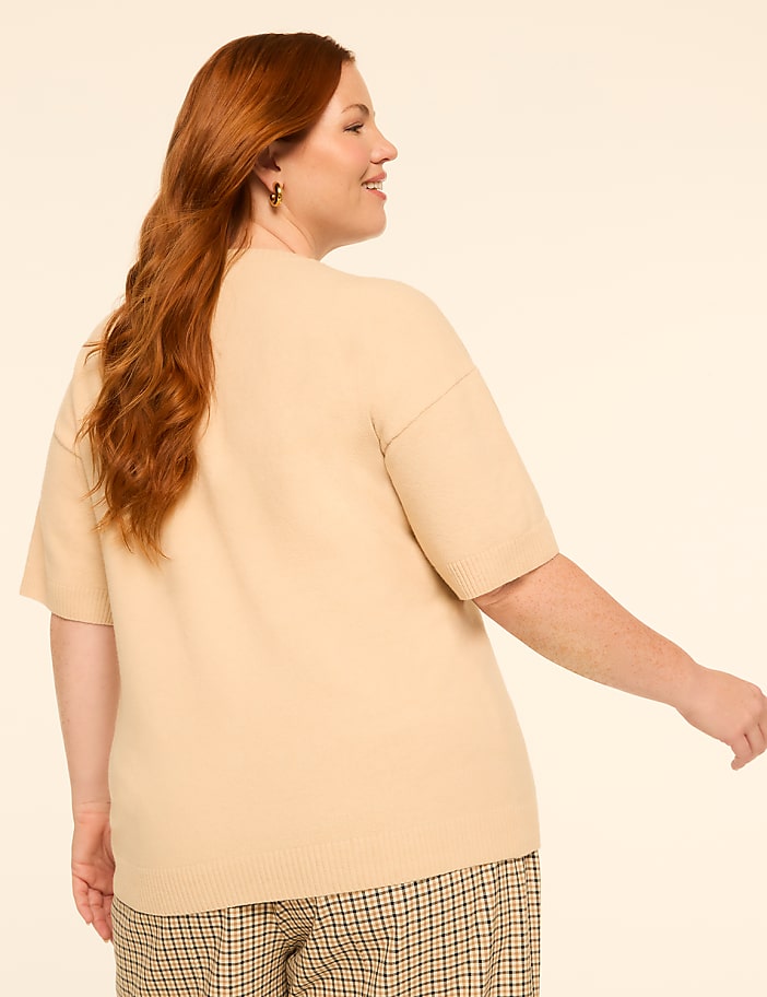 A woman with long red hair wearing a beige sweater and plaid pants turns away from the camera against a plain, light background.