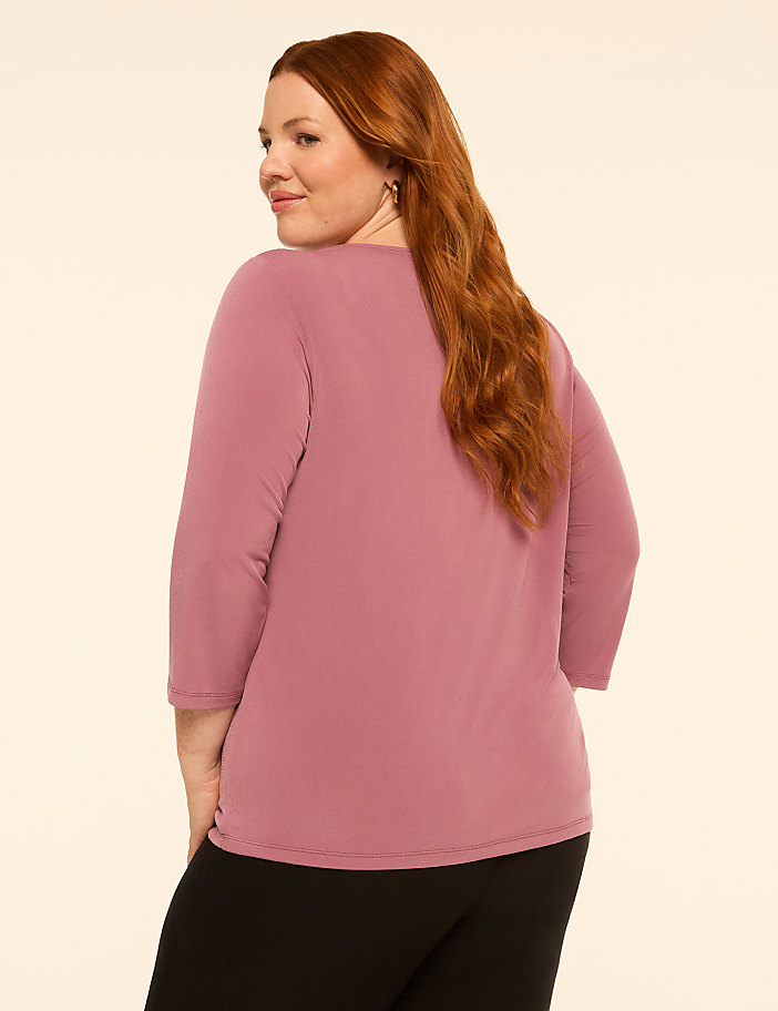 A woman with red hair wearing a mauve top and black pants stands against a plain, light-colored background.